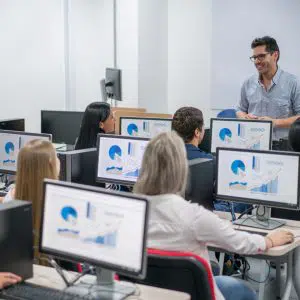 A smiling instructor engaging with employees in a modern computer lab classroom, where each employee is equipped with a desktop displaying graphs and charts for continuing education.